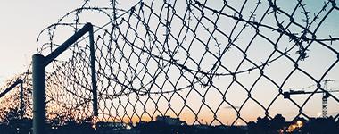 close-up image of barbed wire across a chain link fence with a sunset in the background